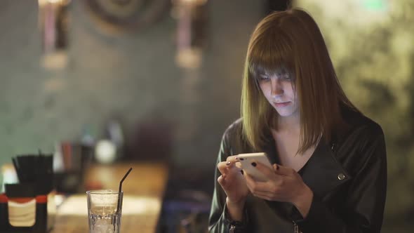 Young Beautiful Redhaired Woman Sitting in Cafe or Bar and Using a Smartphone