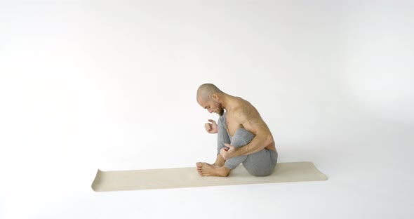 Bald Man with a Bare Torso Performs a Yoga Exercise on a Gym Mat in a White Isolated Room