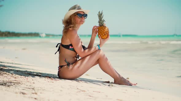 Tanned Girl In Bikini Sitting On Sand. Sexy Blonde Travel Girl Relaxing On Caribbean Beach.
