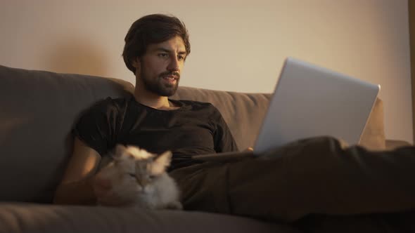 Man Working on a Laptop at Home While Sitting on the Couch and Petting a Cat
