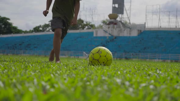 Asian Boy Running And Kicking Football Lying On Green Grass alt