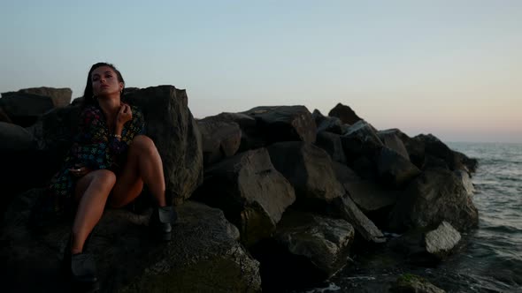 a Brunette in a Swimsuit Shirt and Rough Shoes is Sitting on Large Rocks on the Seashore