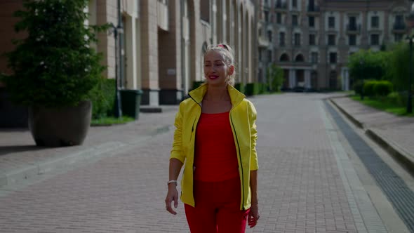a Woman in Red Clothes and a Yellow Jacket Smiles and Walks Along the City Buildings