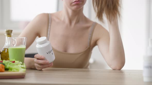 Slim Young Unrecognizable Woman Sitting at Table with Green Smoothie Holding Vitamin E Bottle