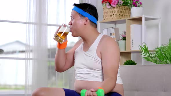 Asian man wearing sport outfits sitting, doing dumbbell and drinking tea at home