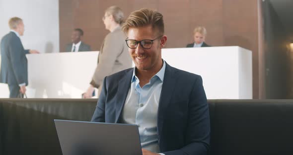 Happy Young Businessman Working on Laptop and Smiling Sitting at Office Building Lobby