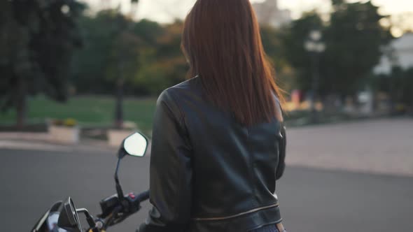 Stylish Young Woman Motorcyclist in a Leather Jacket Goes to Her Bike and Puts on a Helmet