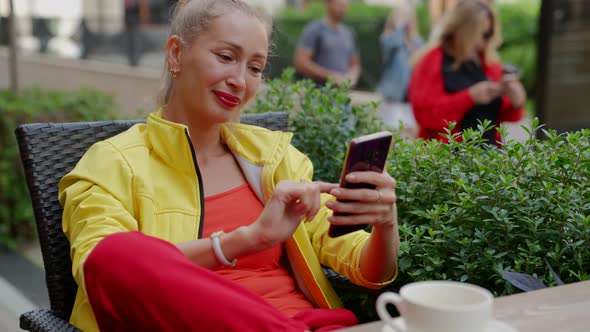 a Blonde in Red and Yellow Clothes Sits with a Phone in Hands and Pokes at the Screen at a Table in