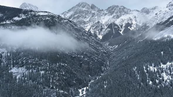 Drone flies up to cloud descending to the slopes of winter mountains in Kananaskis, Alberta, Canada