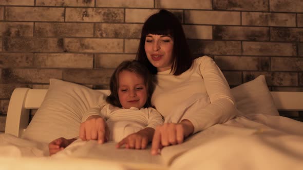 Cute Girl Lying Under the Blanket in Bed Reading Fairytale Story Book with Her Lovely Mother