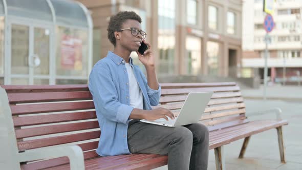 Young African Man Talking on Phone and Using Laptop While Sitting Outdoor on Bench