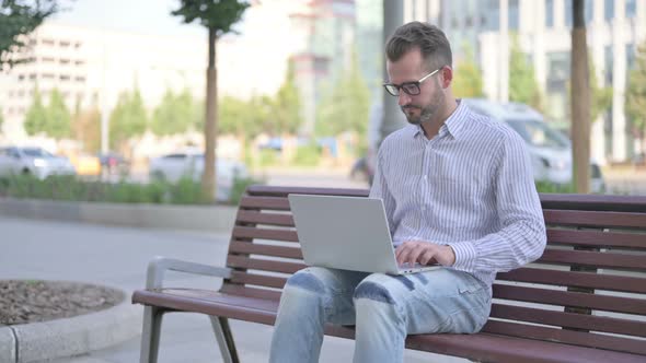 Young Adult Man with Laptop Showing Thumbs Up Sign While Sitting Outdoor on Bench