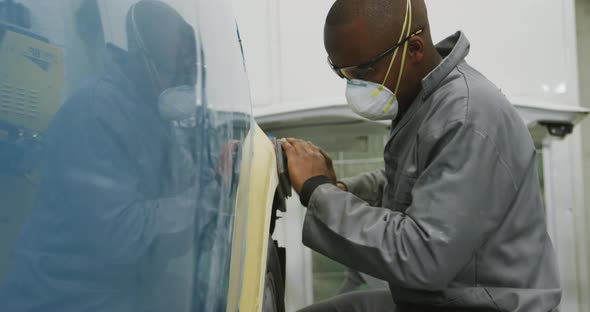 African American male car mechanic wearing a face mask and using a polisher on the side of a car