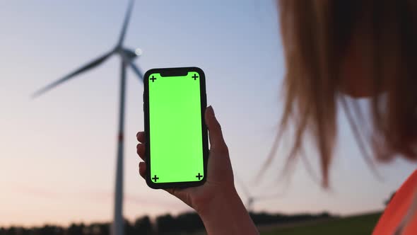 Woman Engineer Holding a Smartphone with Chroma Key on the Background of Wind Turbine at Sunset