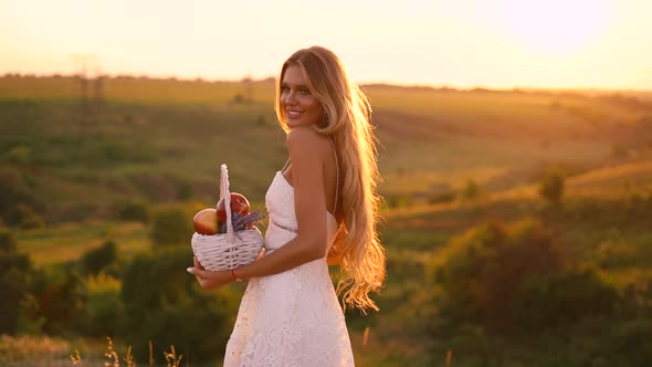 Beautiful sexy blonde girl in white dress posing in a field at sunset with a basket of fruit	