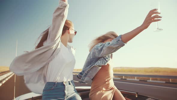 Two Beautiful Young Women Fun Ride in Convertible Raising Their Hands Up with Champagne