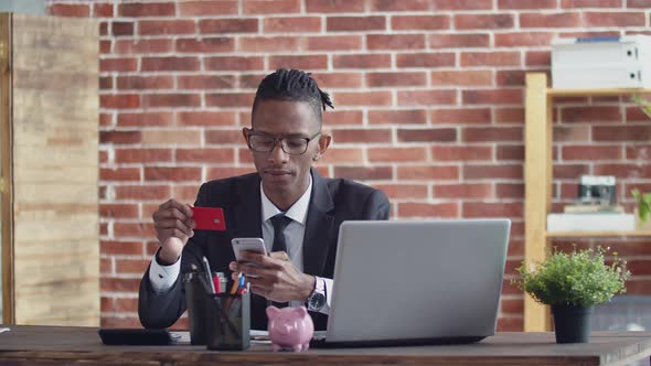 Serious Black Man in Glasses and a Strict Suit Enters the Bank Card Data Into Phone Sitting at an