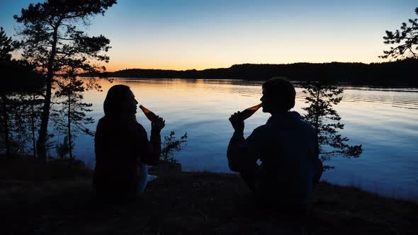 Couple Clinks Bottles of Beer Sitting on Bank of Calm River