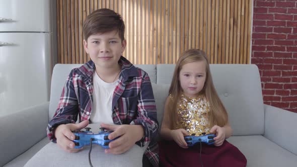 Portrait of Cute Caucasian Girl and Boy Sitting on Sofa and Playing Video Game. Smiling Children