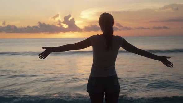 Backside View, Silhouette of Woman Looks at Ocean, Sunset and Spreads Arms Wide