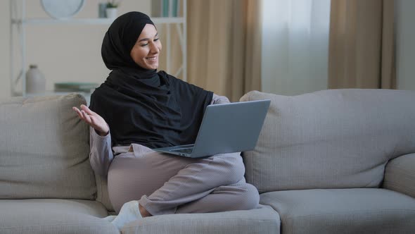 Friendly Arabian Muslim Woman Girl in Hijab Sit on Cozy Sofa Talk Video Call with Friend Greeting at