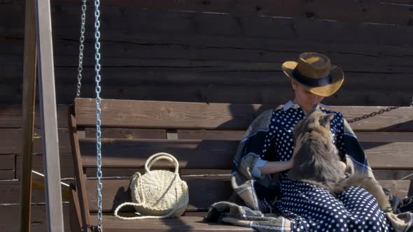 Girl in a Hat Resting on a Wooden Swing in the Village with a Fluffy Cat