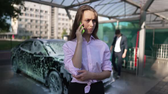 Serious Young Caucasian Woman Talking on Phone As Man Washing Car at Background at Service Station