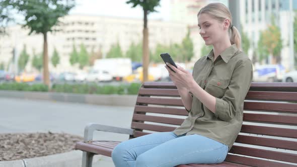 Woman Celebrating Online Success on Smartphone while Sitting Outdoor on Bench