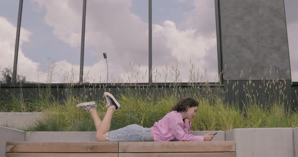 Happy European teen having fun and lying on the bus stop with a smartphone.