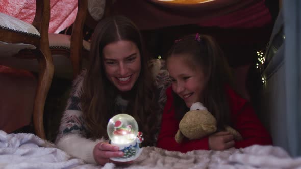 Caucasian mother and daughter holding snow globe while lying under blanket fort during christmas