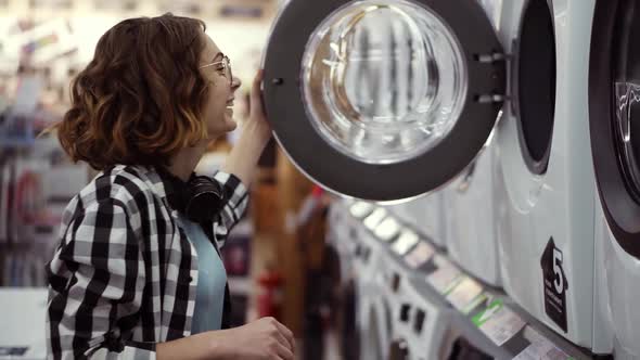 A Young Positive Woman in a Plaid Shirt Choosing Washing Machine in the Shop of Household Appliances