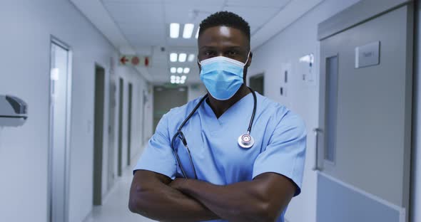 Portrait of african american male doctor wearing face mask and scrubs standing in hospital corridor