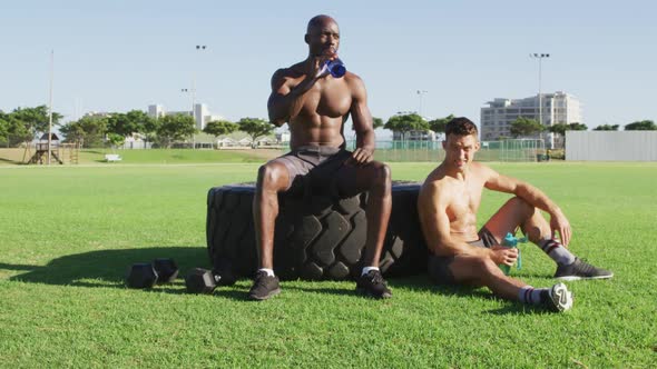 Two fit, shirtless diverse men cross resting, drinking water after exercising outdoors