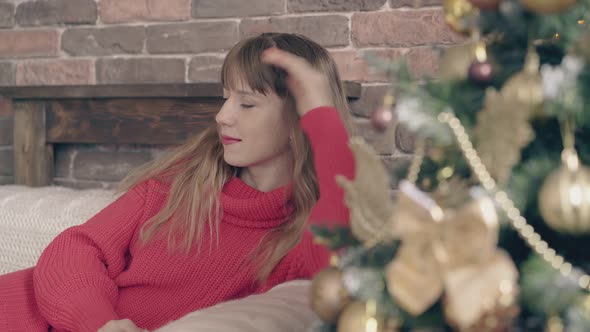 Young Fair-haired Girl Dreams on Couch Near Christmas Tree