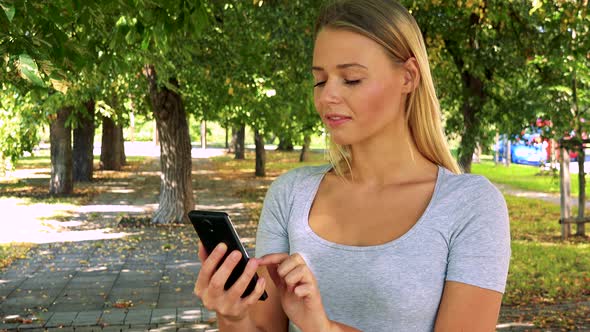 Young Pretty Blond Woman Works (Typing) on Smartphone - Park with Trees in Background