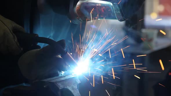 A man wearing a welding helmet and gloves uses an arc welder on a silver metal arrowhead held in a v
