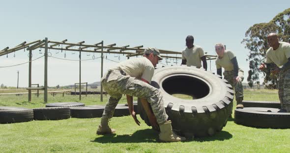 Fit caucasian male soldier flipping tractor tyre, with diverse group at army obstacle course