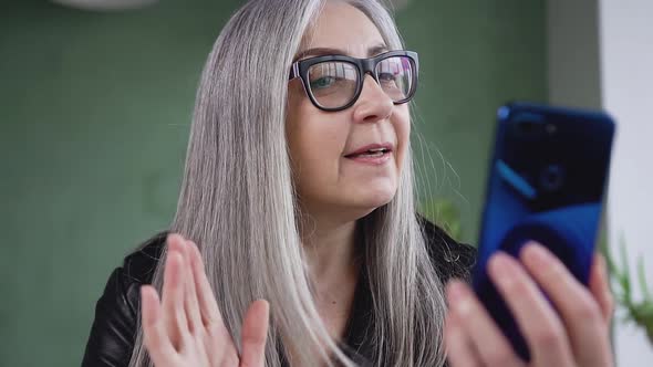 Happy Senior Woman in Glasses with Long Gray Hair which Having Video Call Using Blue Phone