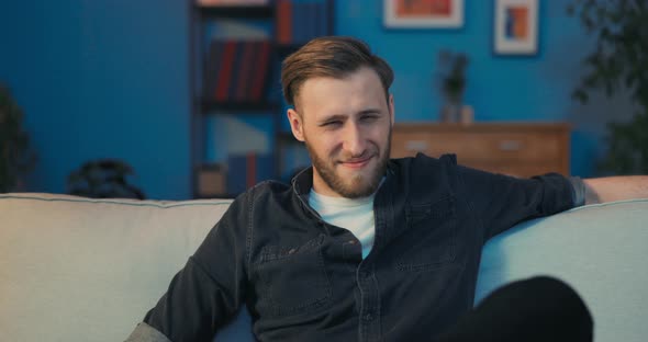 A Handsome Man is Sitting on Sofa Late at Night in Front of TV Screen