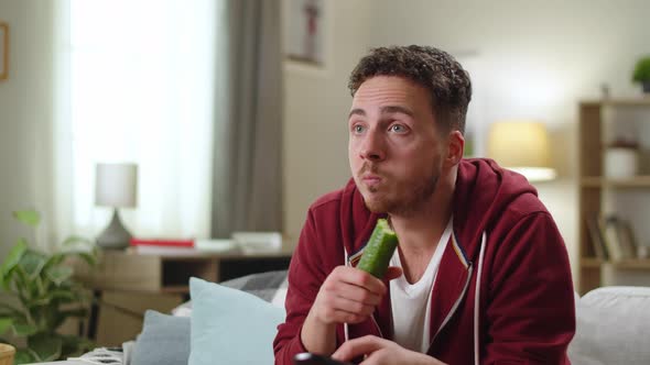 Man Sitting at Home on the Couch and Biting a Cucumber