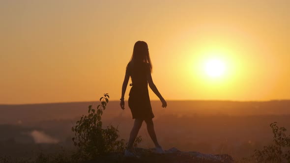 Dark Silhouette of a Young Woman Jumping with Raised Up Hands on a Stone Enjoying Sunset View