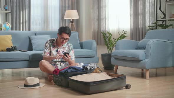 Asian Young Man Using Smart Phone While Packing Clothes In Suitcase At Home, Preparing For Vacation