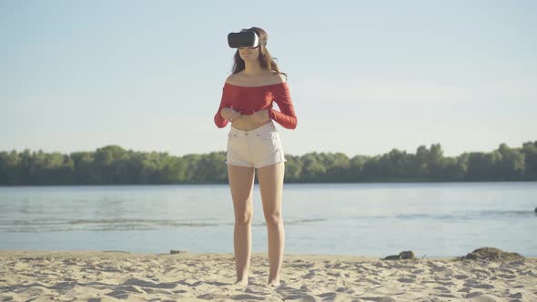 Wide Shot of Young Slim Brunette Woman Standing on River Bank in VR-googles and Looking Around