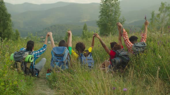 Cheering Multiethnic Tourists with Backpacks Sitting with Arms Raised on Mountain Hill