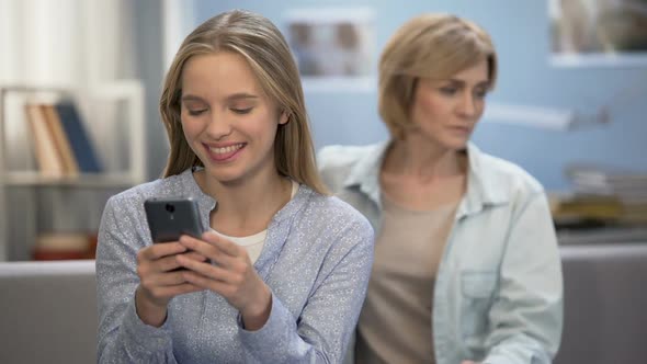 Happy Teenage Girl Texting Messages in Smartphone, Sad Mother Sitting on Sofa