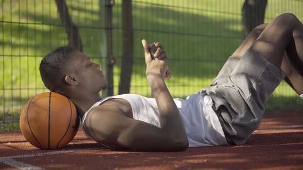 Side View of Young African American Man Lying on Ball and Using Smartphone. Portrait of Positive