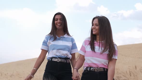 Front view of two female friends holding hands and smiling while walking in a wheat field together.