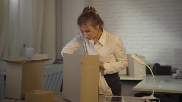 Young Confident Woman Packing Cardboard Box and Tapping on Tablet. Portrait of Caucasian Seamstress