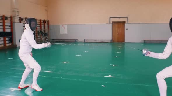 Two Young Women in White Protective Suits Having a Fencing Training in the School Gym