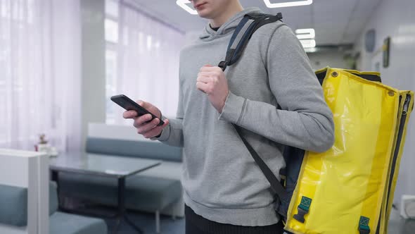 Unrecognizable Male Courier Checking Order in Smartphone App Standing in Cafe with Yellow Thermal
